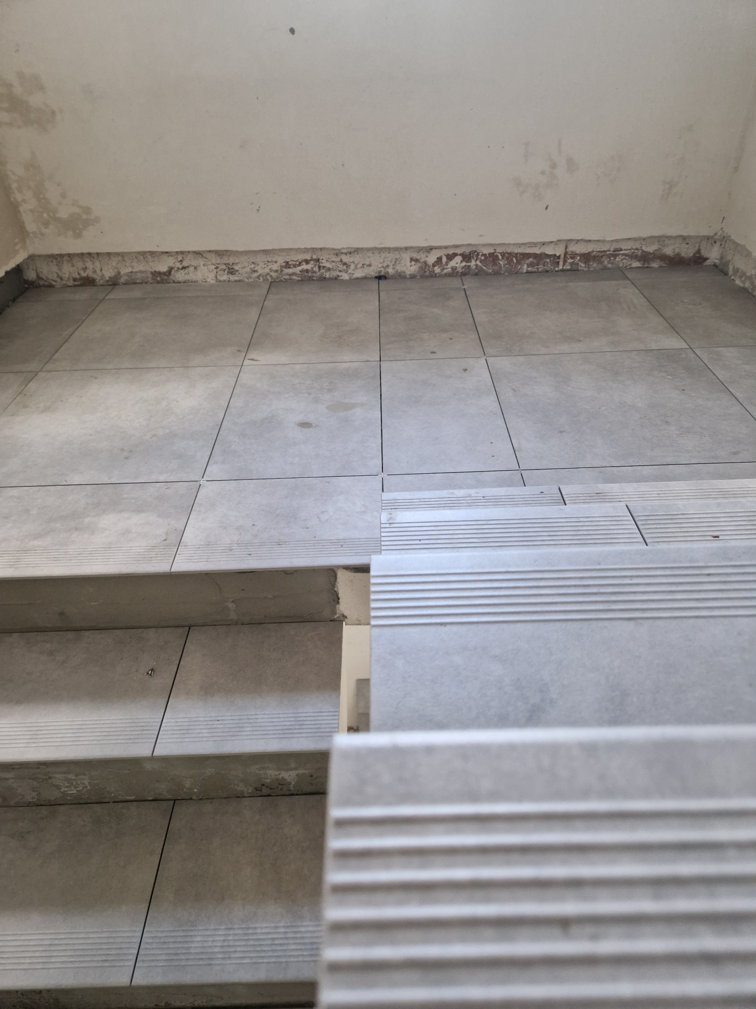 Szare płytki podłogowe i schody z wykończeniem antypoślizgowym, widoczne ślady prac remontowych na ścianie przy podłodze.