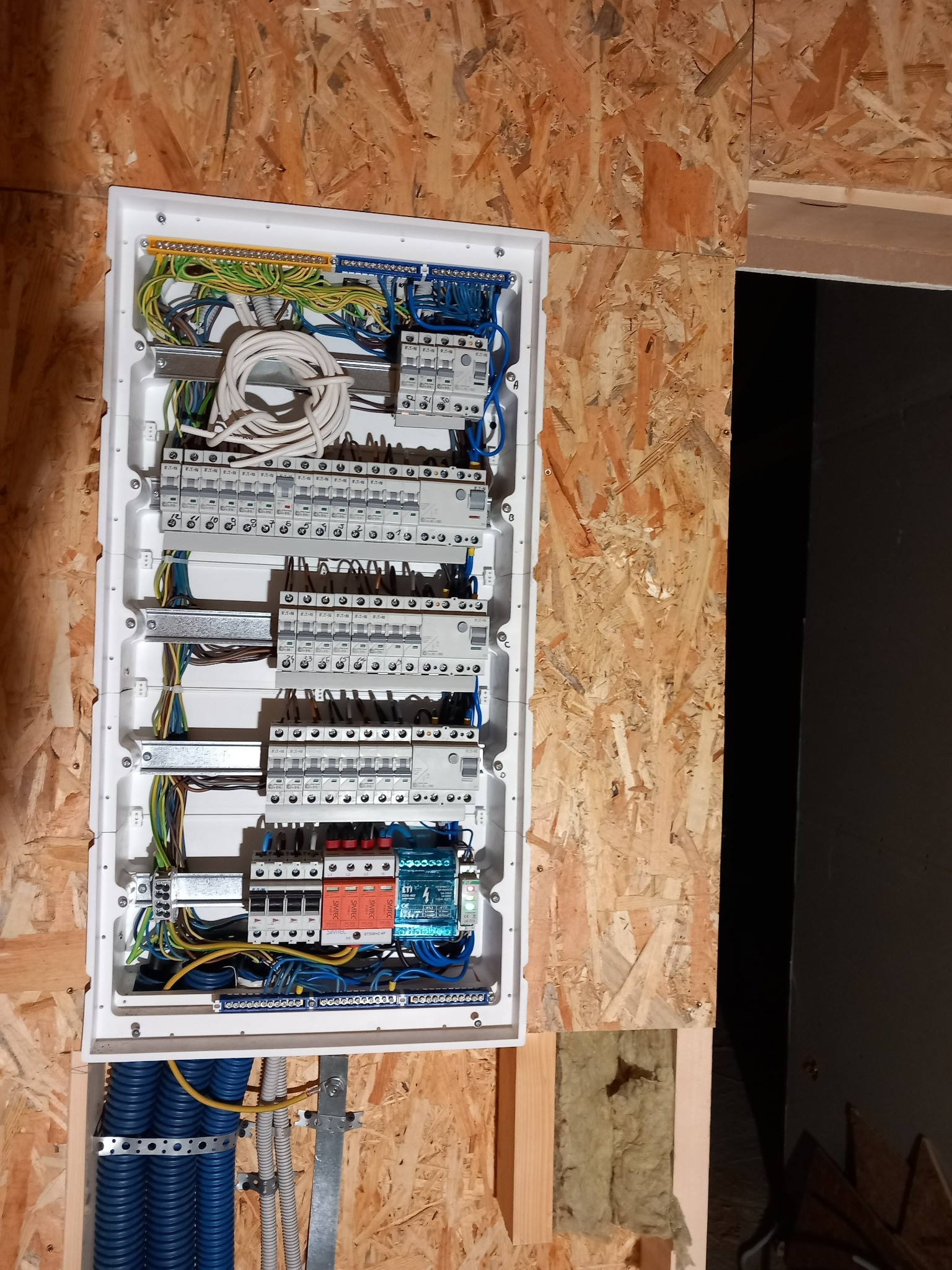 Rozdzielnica elektryczna w obudowie, zamontowana na ścianie z płyty OSB, z widocznymi przewodami, bezpiecznikami i zabezpieczeniami przepięciowymi.