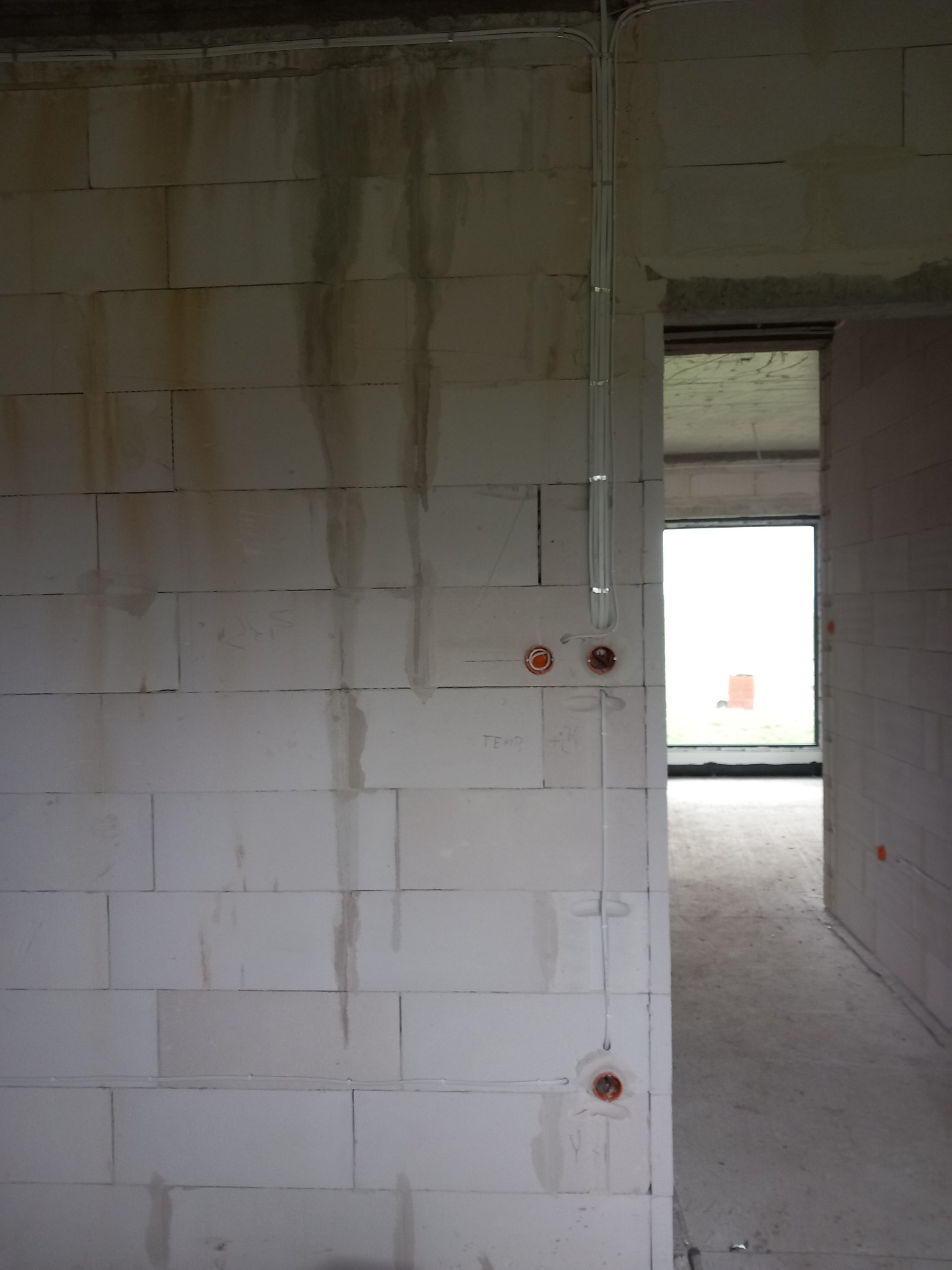 Surowe wnętrze w trakcie budowy z widocznymi przewodami elektrycznymi i puszkami instalacyjnymi na ścianie z betonu komórkowego.