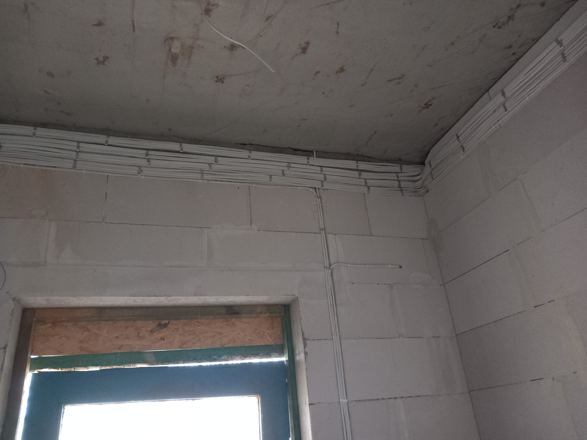 Białe przewody elektryczne ułożone w korytkach na suficie i ścianie z betonu komórkowego w surowym stanie, widoczne okno z częściowo zamontowaną ramą.