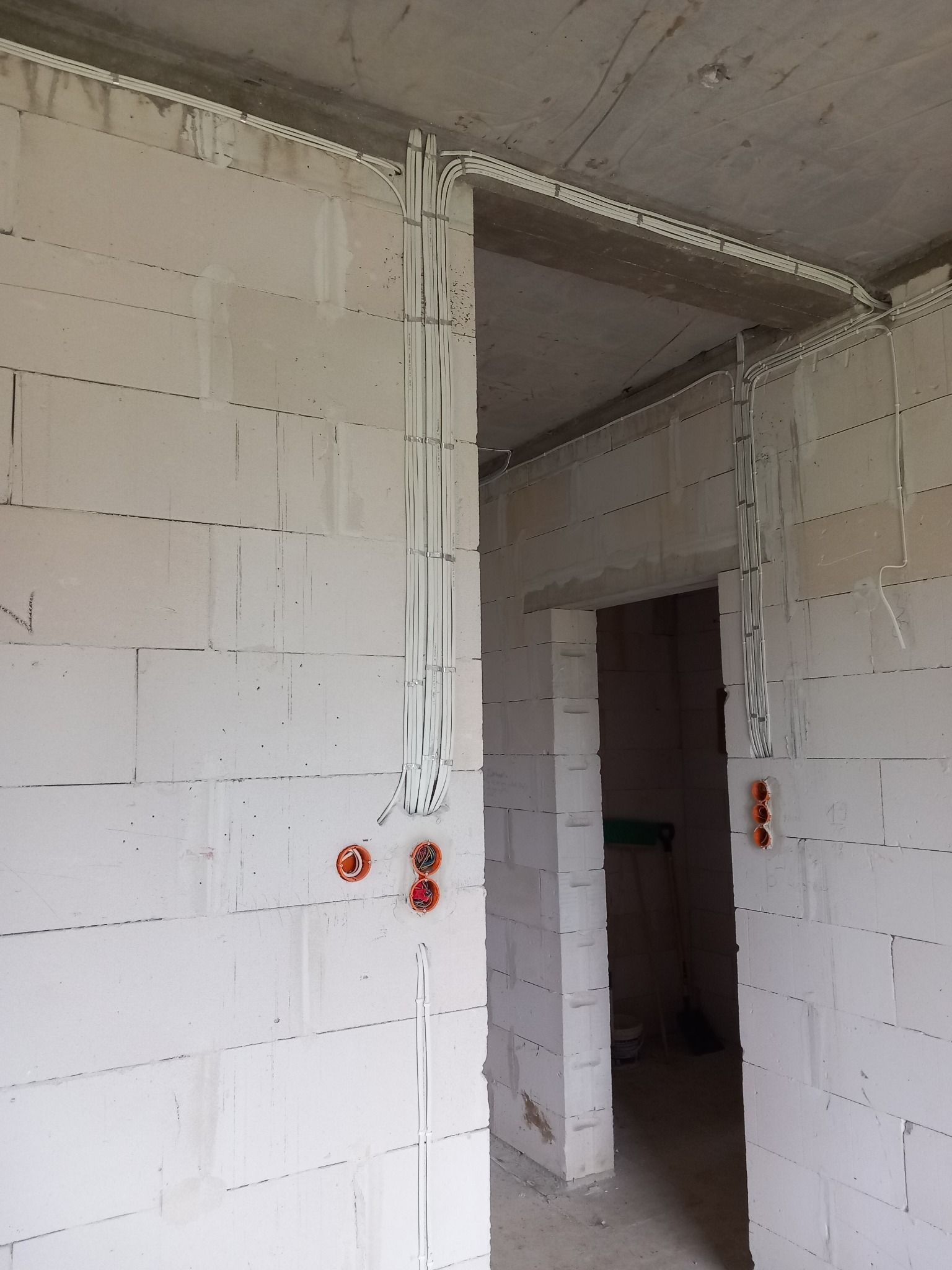 Surowe wnętrze w trakcie prac wykończeniowych. Widoczne białe ściany z betonu komórkowego, instalacja elektryczna w białych rurkach prowadzona pod sufitem i pionowo wzdłuż ścian, pomarańczowe...