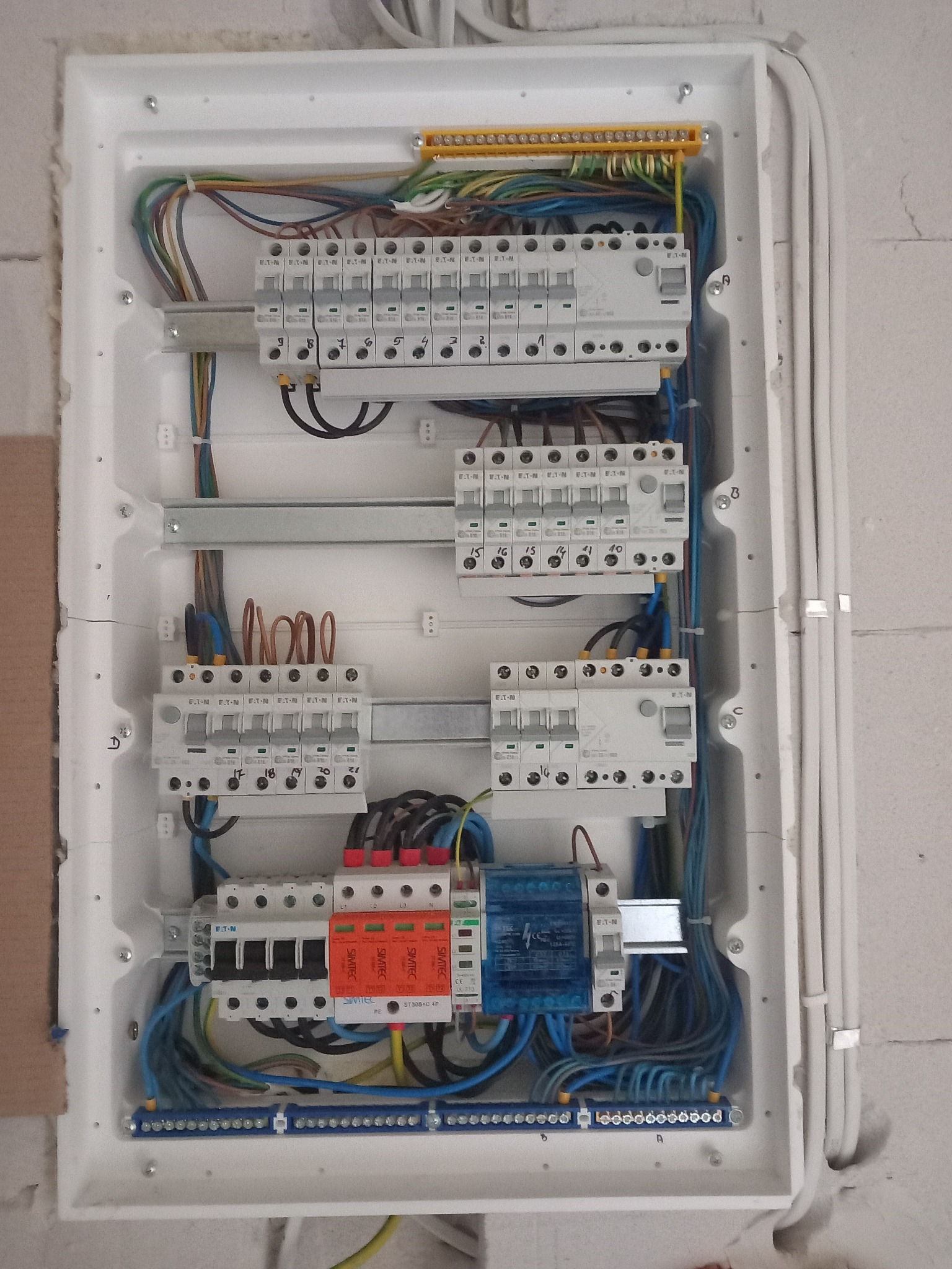 Otwarta skrzynka rozdzielcza z bezpiecznikami, zabezpieczeniami przepięciowymi i przewodami w różnych kolorach, zamontowana na surowej ścianie.