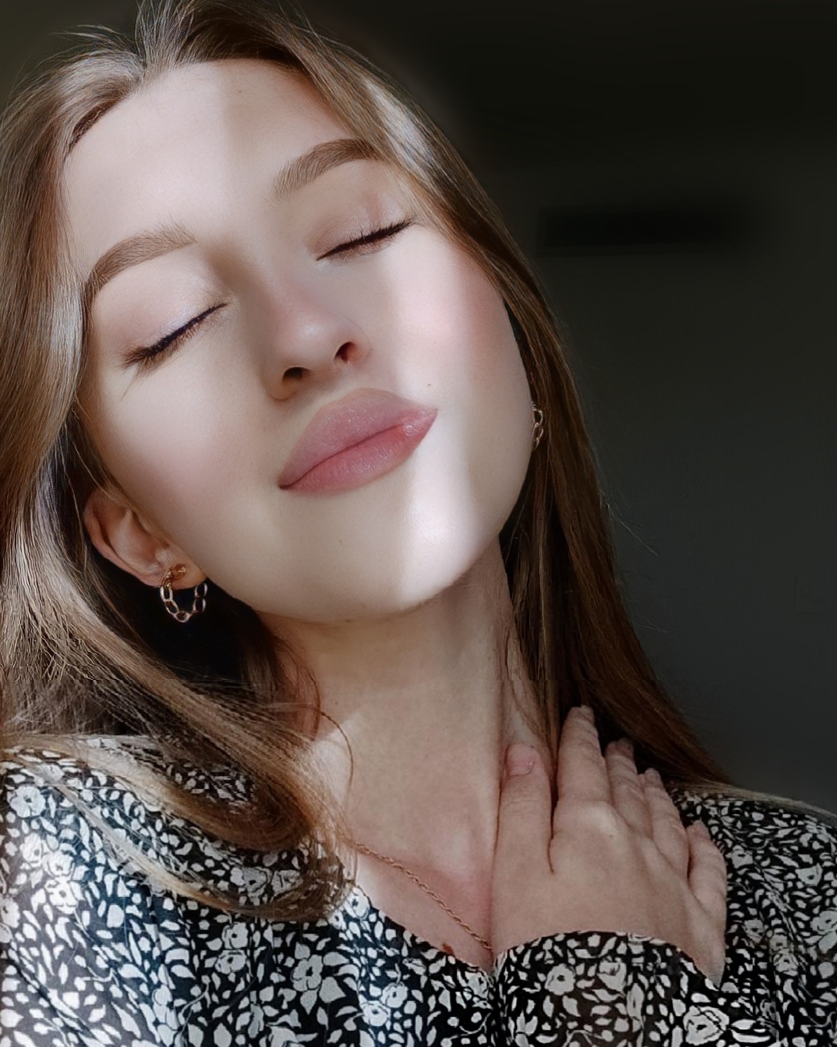 Portret kobiety z zamkniętymi oczami, z ręką na szyi, w sukience w kwiaty, oświetlonej kontrastowym światłem.