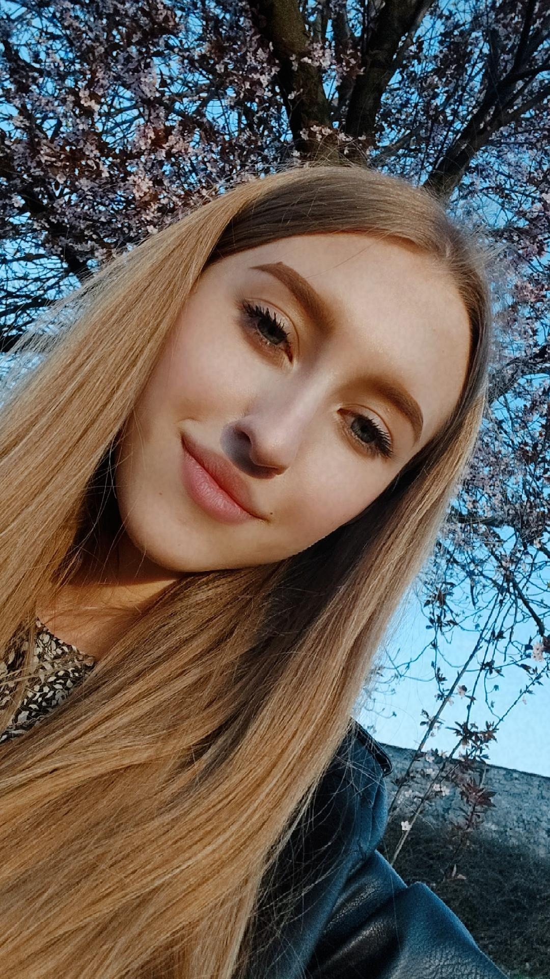 Portret młodej kobiety z długimi blond włosami na tle kwitnącego drzewa i błękitnego nieba.