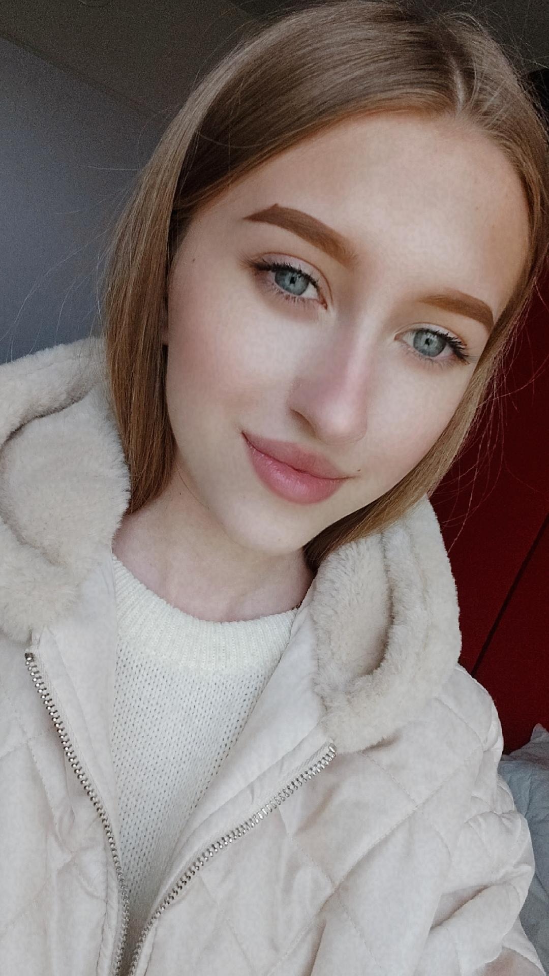 Portret młodej kobiety z jasnymi włosami i niebieskimi oczami, ubrana w biały sweter i kurtkę z futrzanym kołnierzem, z delikatnym makijażem i subtelnym uśmiechem.