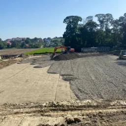 Pomarańczowa koparka rozładowuje żwir na przygotowany teren z widocznymi śladami opon, w tle budynki mieszkalne i drzewa.