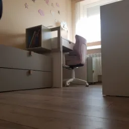 Pokój dziecięcy z drewnianą podłogą, biurkiem z szufladami i półką na książki, różowym krzesłem obrotowym oraz dekoracjami ściennymi w kształcie chmur, gwiazd i liter