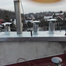 Komplet trzech metalowych kominów wentylacyjnych z daszkami, oraz jeden wysoki komin stalowy, zamontowane na szczycie komina murowanego na dachu z czerwonej blachy trapezowej, z widokiem...