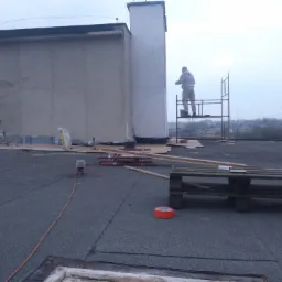 Pracownik na rusztowaniu maluje komin na dachu pokrytym papą, w tle drabina i narzędzia, pochmurny dzień.