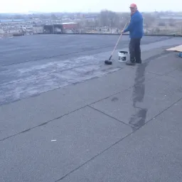 Pracownik w czerwonej czapce maluje płaski dach z papy za pomocą wałka na długim kiju, widoczny kubeł z czarną substancją, w tle zabudowania przemysłowe i drzewa.