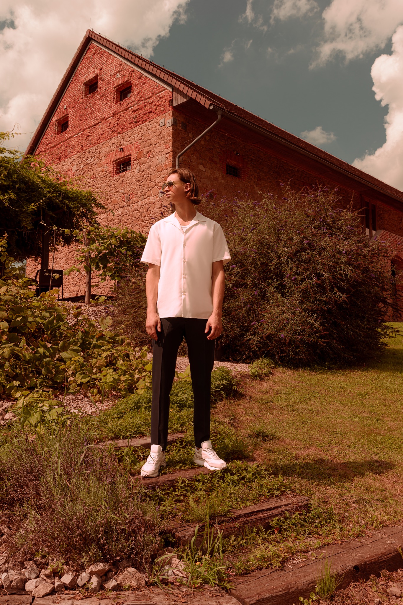 Mężczyzna w białej koszuli i czarnych spodniach pozuje na tle ceglanego budynku i zieleni. Ubrany w okulary przeciwsłoneczne, stoi na drewnianych stopniach pośród roślinności.