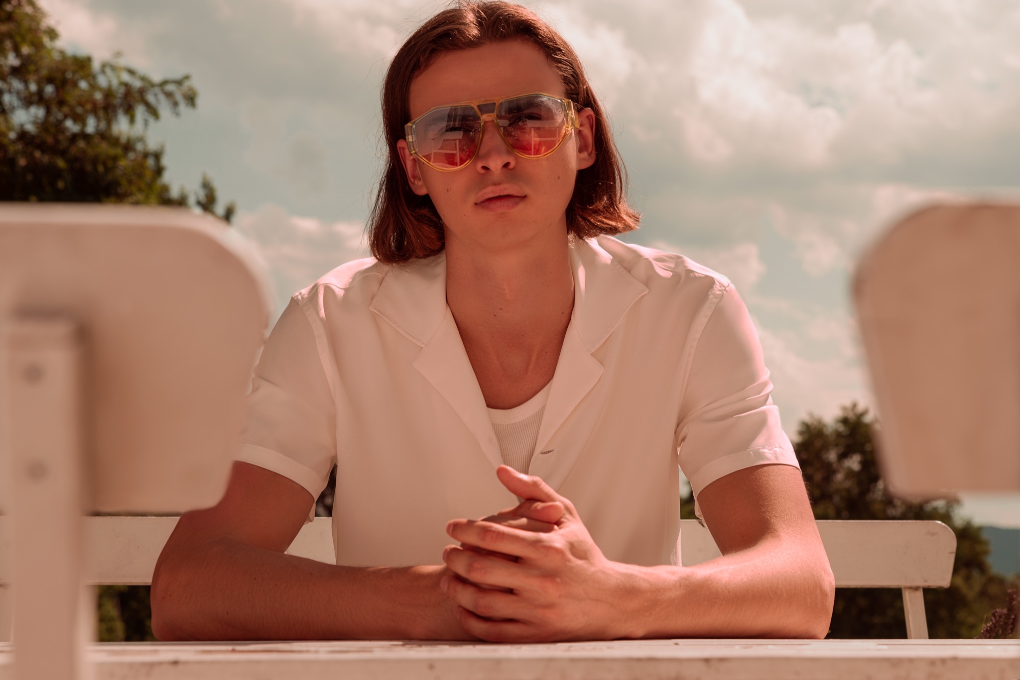 Młody mężczyzna z długimi włosami siedzi przy białym stole na zewnątrz, nosi białą koszulę i duże, żółte okulary przeciwsłoneczne z gradientem kolorów, tło to niebo i drzewa.