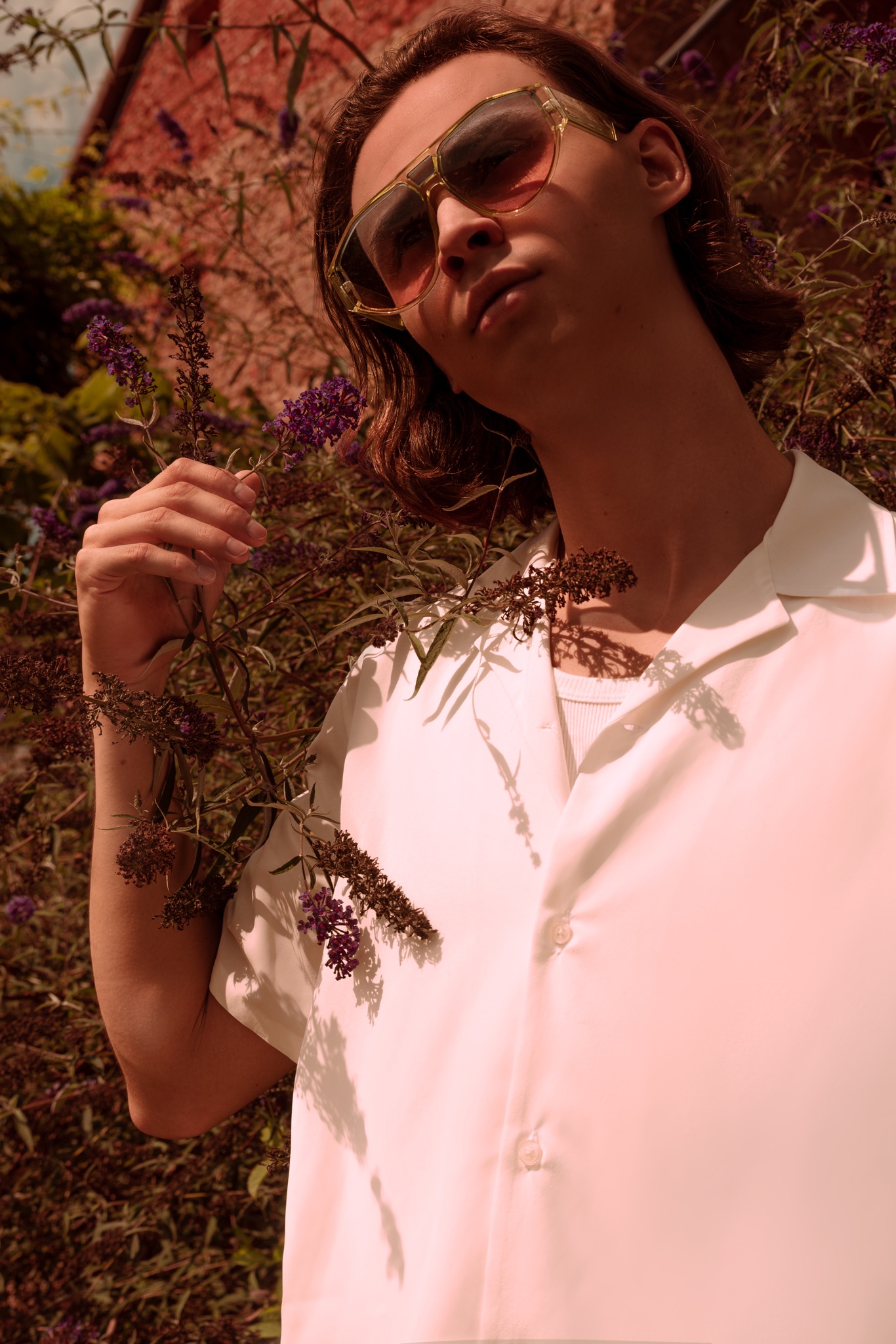 Młody mężczyzna w okularach z żółtymi oprawkami, ubrany w białą koszulę z krótkim rękawem, pozuje na tle kwitnącego krzewu z fioletowymi kwiatami.