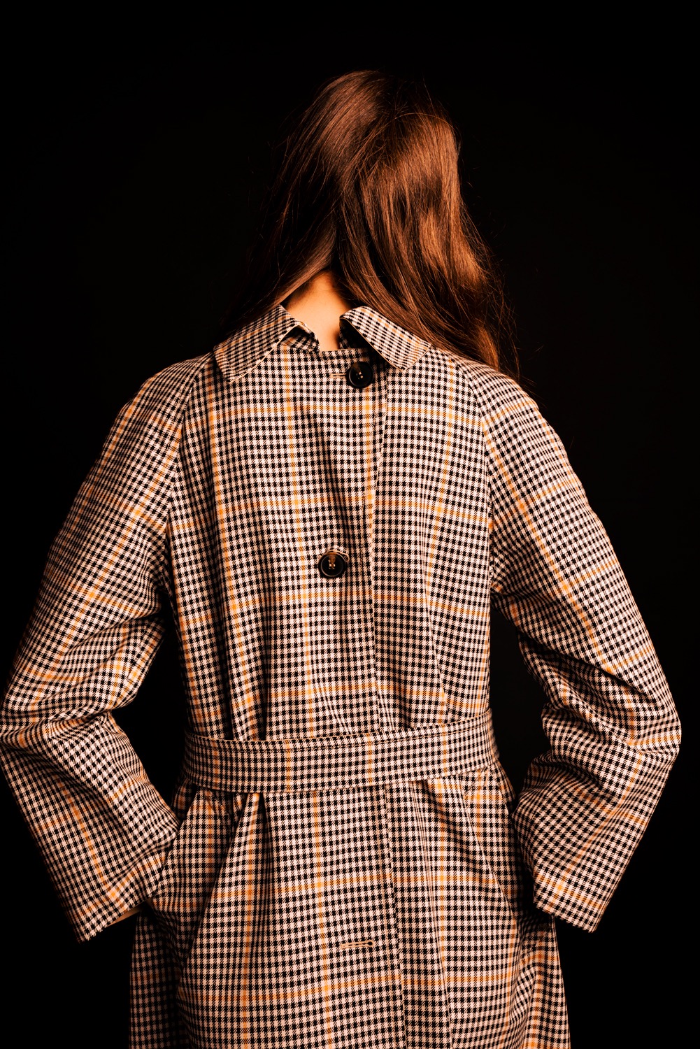 Kobieta odwrócona tyłem, ubrana w płaszcz w kratę z paskiem, na czarnym tle, widoczne detale takie jak guziki i faktura materiału.