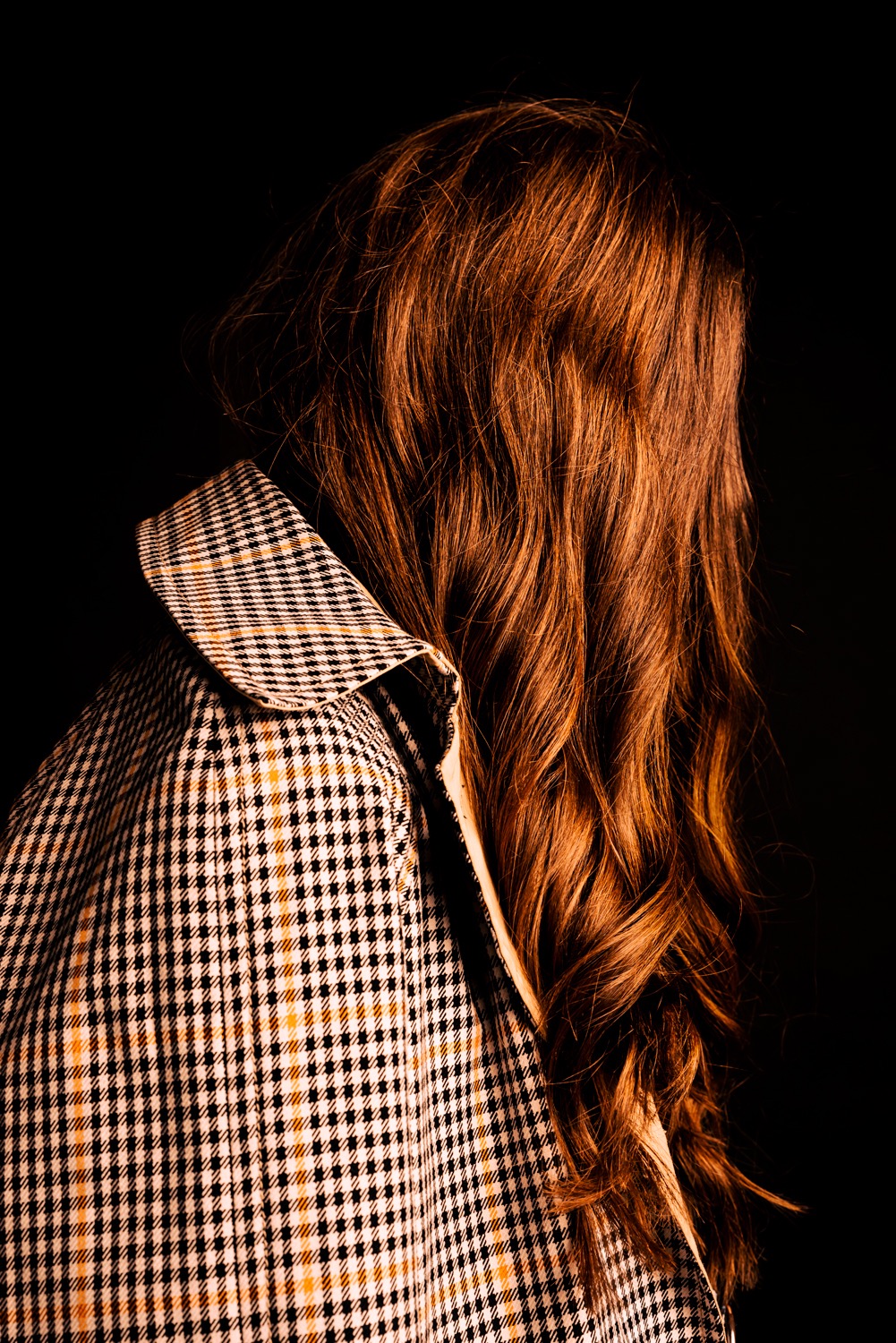 Kobieta z długimi, rudymi, falowanymi włosami, ubrana w płaszcz w drobną kratę na czarnym tle.