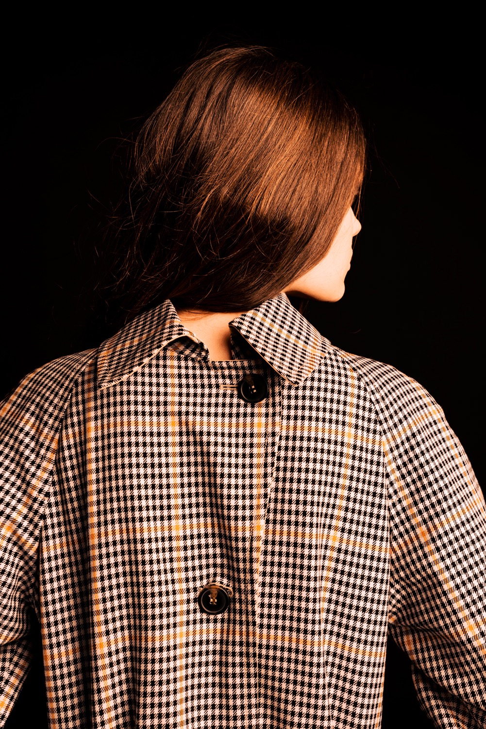 Kobieta odwrócona tyłem, ubrana w płaszcz w drobną kratę z dwoma guzikami na plecach i rozpuszczonymi, brązowymi włosami na ciemnym tle.