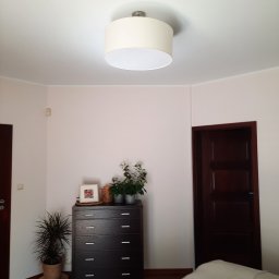 Mońka Piotr - Jasne wnętrze pokoju z kremowymi ścianami, drewnianą podłogą, ciemnymi drzwiami, komodą i roślinami; sufit z okrągłą lampą.