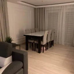 Jasne drewniane panele podłogowe w salonie z szarym narożnikiem, stołem z krzesłami i zasłonami w tle. Pod stołem leży różowy balon.