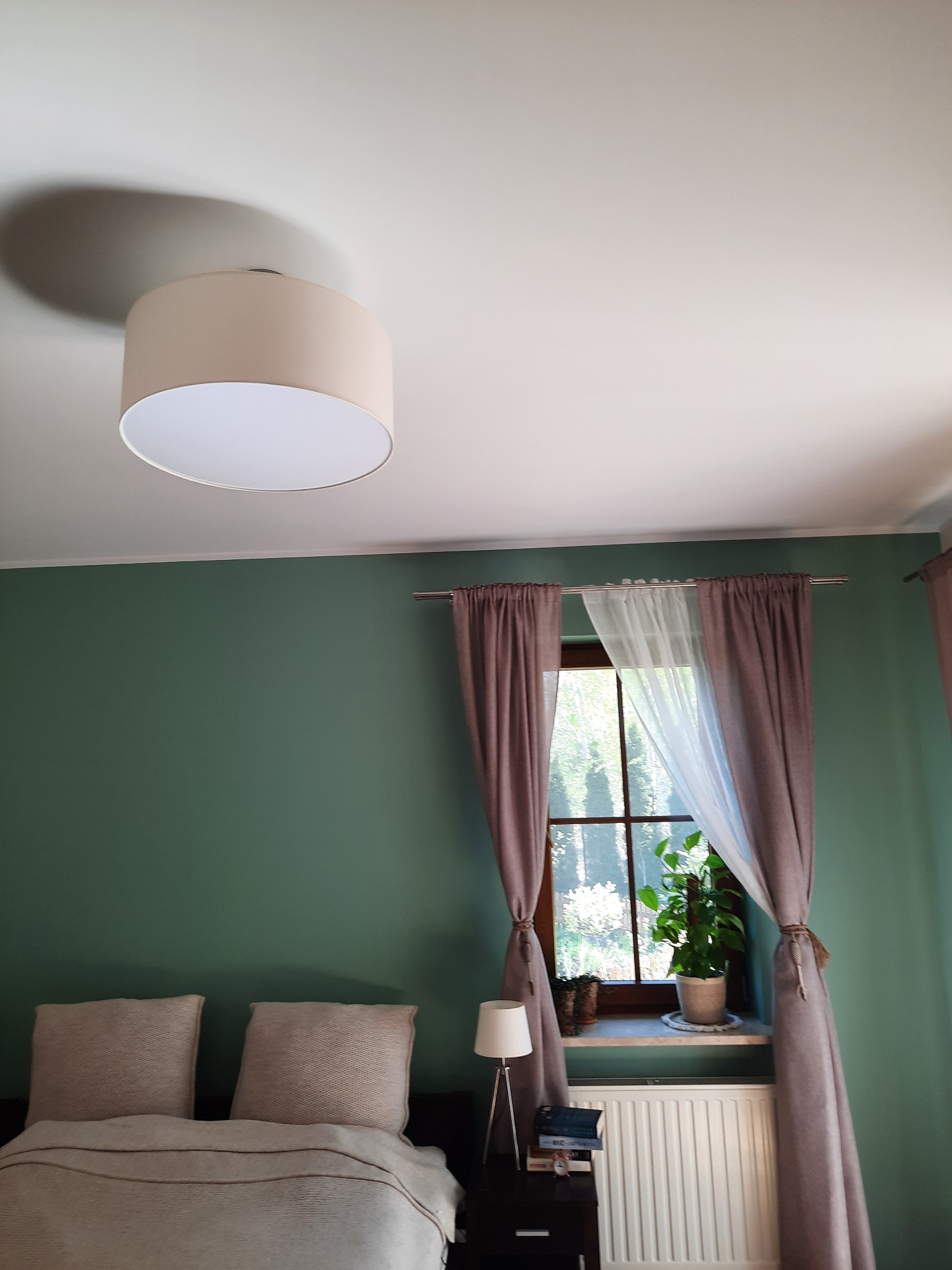 Pokój z zielonymi ścianami, beżowym abażurem lampy sufitowej, oknem z brązowymi i białymi zasłonami, rośliną w doniczce na parapecie i łóżkiem z beżowymi poduszkami.