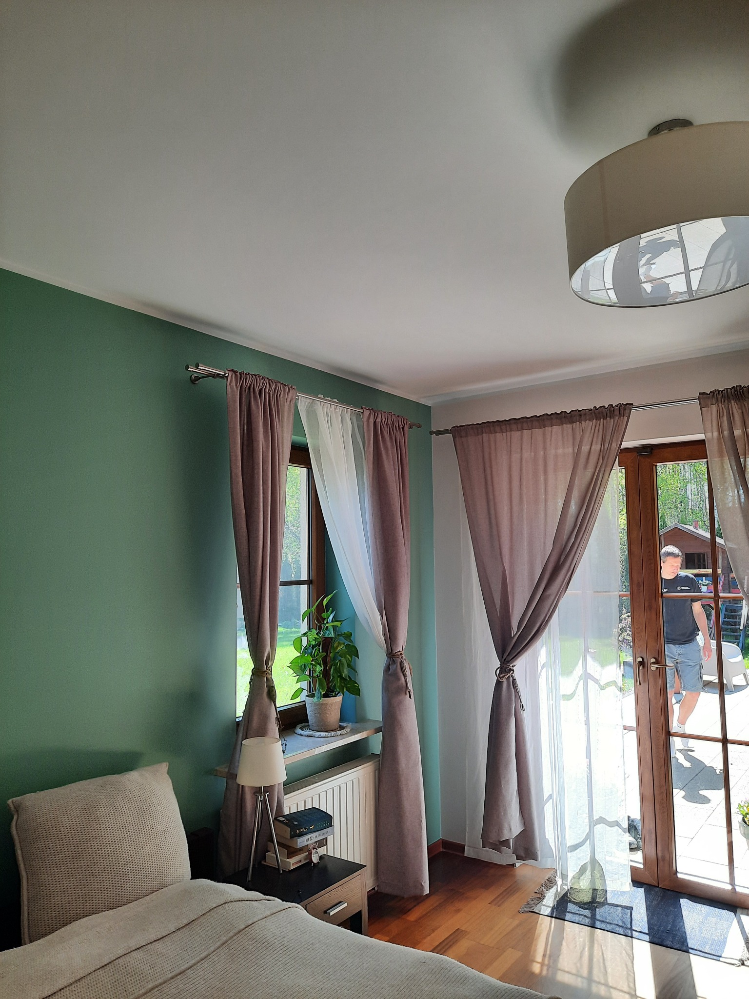 Pokój z zieloną ścianą, drewnianą podłogą, oknem z brązowymi i białymi zasłonami związanymi sznurkiem, oraz wyjściem na taras, przez które widać mężczyznę.