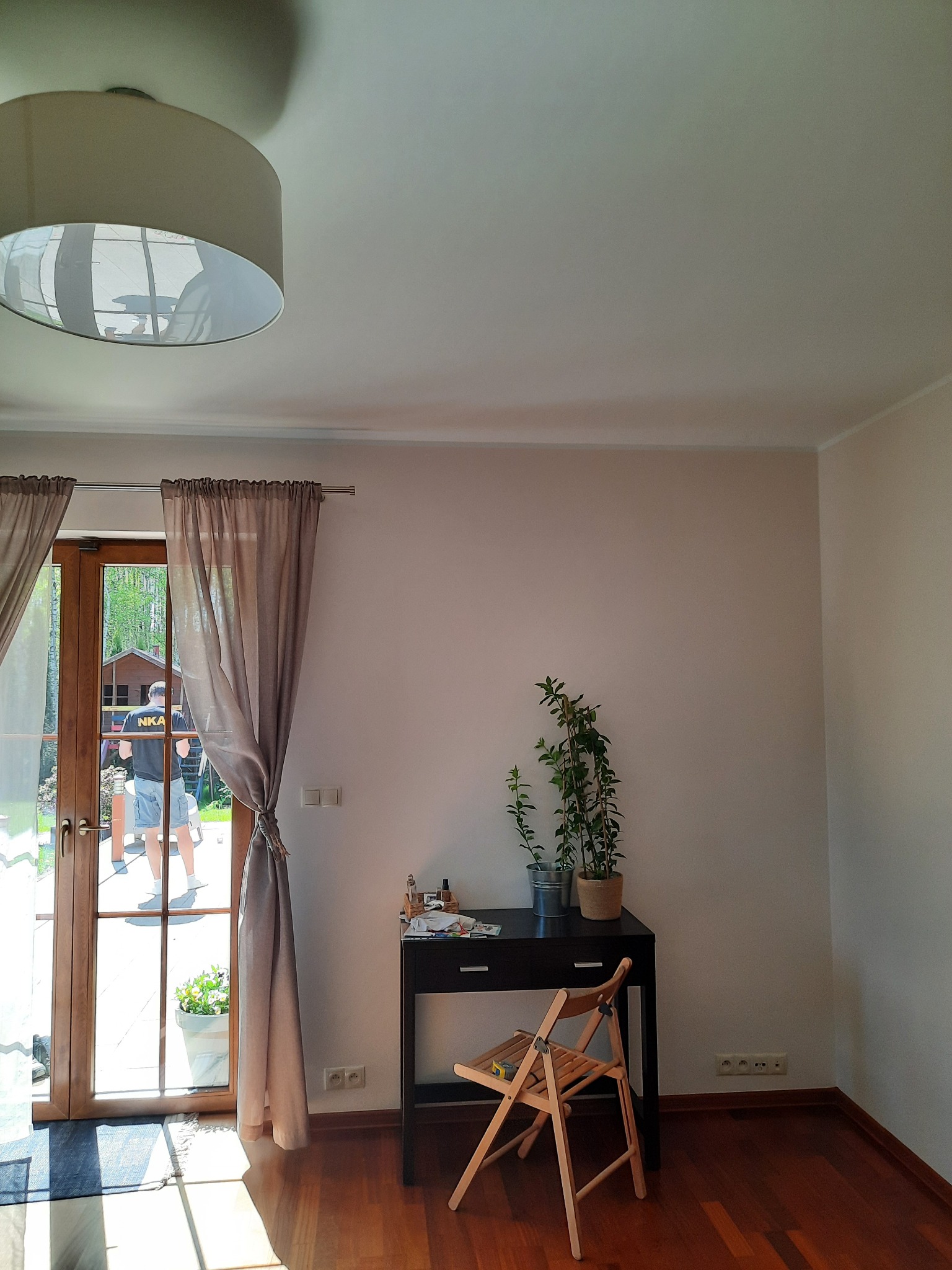 Jasne wnętrze pokoju z drewnianą podłogą, biurkiem z roślinami, krzesłem składanym i oknem balkonowym z zasłonami, przez które widać osobę na zewnątrz.