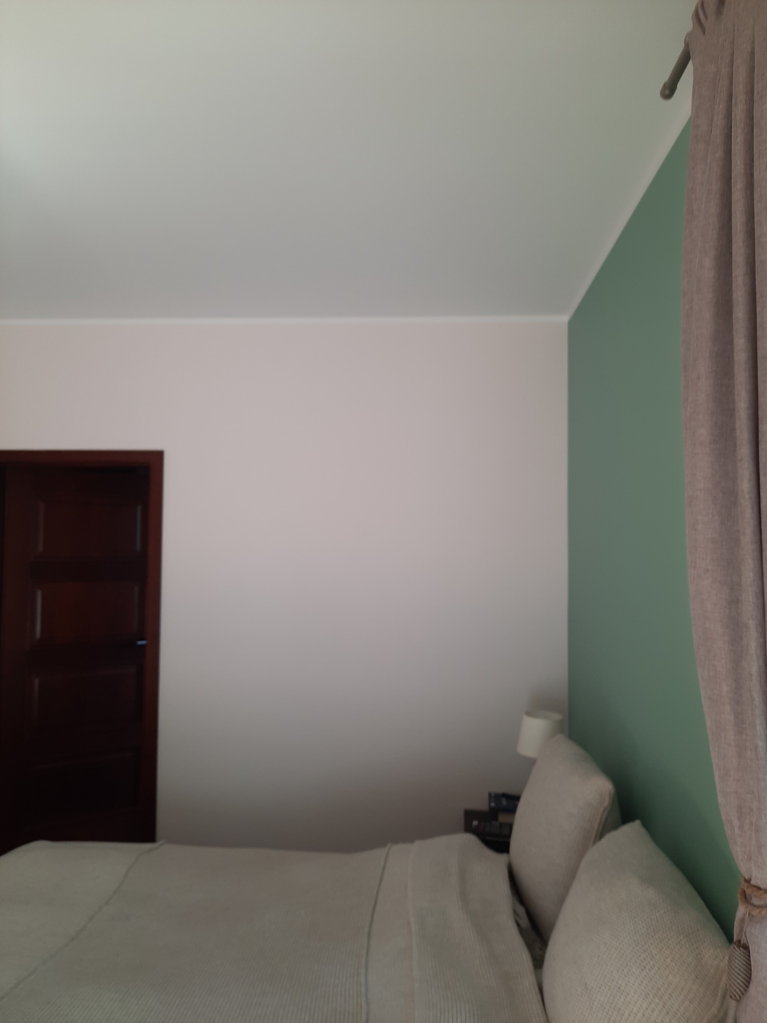 Pokój po odświeżeniu: ściana pomalowana na zielono kontrastuje z kremową ścianą, widoczna drewniana ościeżnica drzwi i fragment łóżka z poduszkami, jasne zasłony z boku.