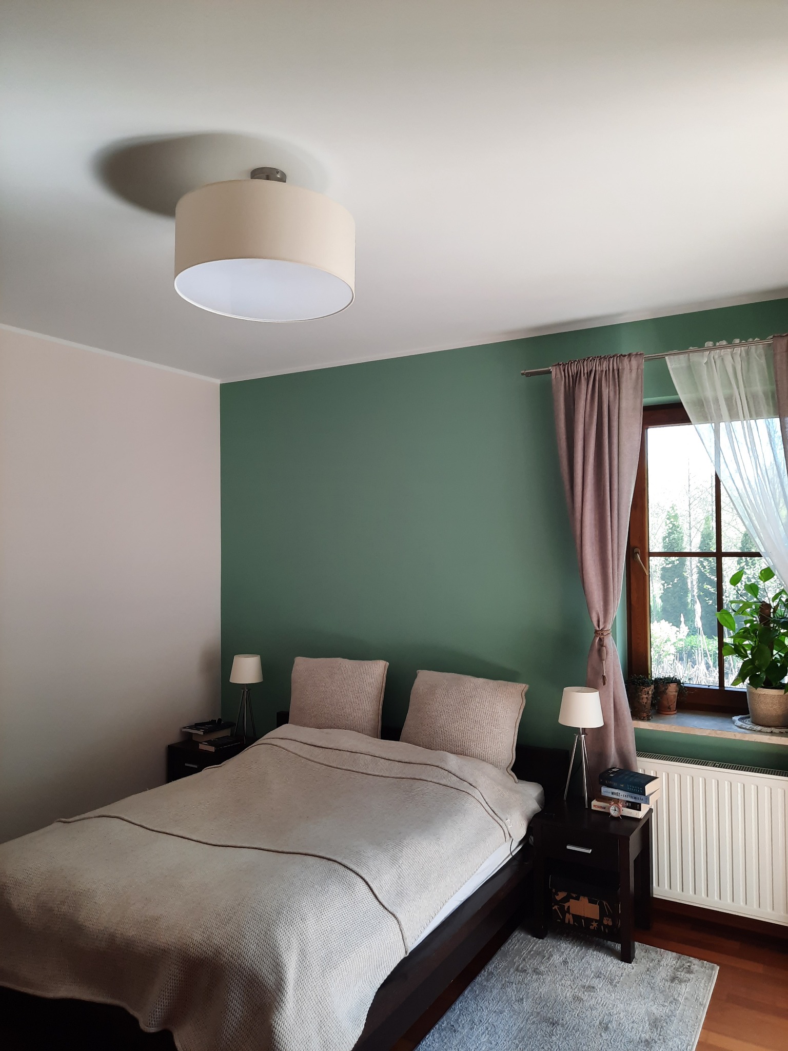Sypialnia z łóżkiem z beżową narzutą, ścianą pomalowaną na zielono i różowo, lampką nocną na stoliku oraz oknem z zasłonami w odcieniach brązu i bieli.