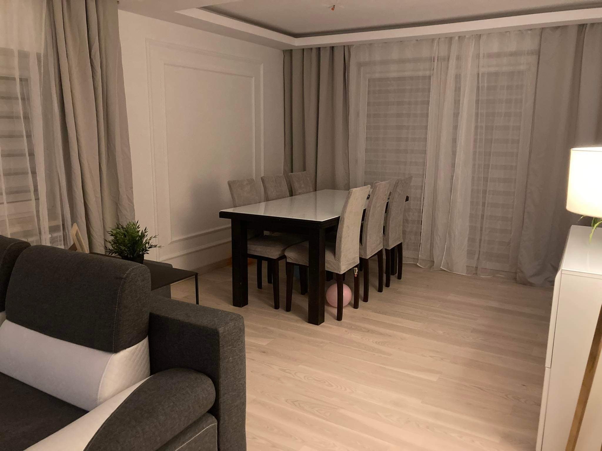 Jasne drewniane panele podłogowe w salonie z szarym narożnikiem, stołem z krzesłami i zasłonami w tle. Pod stołem leży różowy balon.