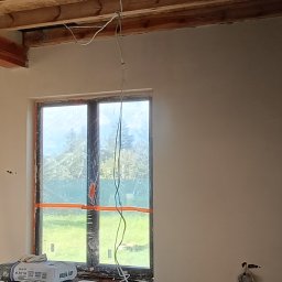 AMBITION Jarosław Rozwadowski - Wnętrze po nałożeniu gładzi, widoczne drewniane belki stropowe, okno zabezpieczone folią, drabina malarska i worki z zaprawą. Ściany gładkie, przygotowane do dalszych prac.