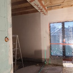 AMBITION Jarosław Rozwadowski - Wnętrze w trakcie prac wykończeniowych. Widoczne ściany po szpachlowaniu, laser krzyżowy wyznaczający poziom, worki z zaprawą i drabina. Surowe drewno na suficie.