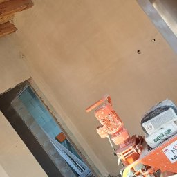 AMBITION Jarosław Rozwadowski - Świeżo otynkowane ściany wewnątrz budynku z widoczną maszyną tynkarską Malek M-4 MAX i workami z zaprawą. Widoczne drewniane elementy konstrukcyjne stropu.