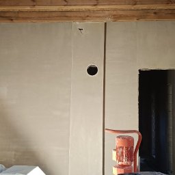 AMBITION Jarosław Rozwadowski - Ściana po nałożeniu tynku maszynowego, widoczne drewniane belki stropowe, otwór w ścianie i maszyna do tynkowania. Świeży tynk o jednolitej fakturze.