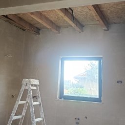 AMBITION Jarosław Rozwadowski - Wnętrze w trakcie prac wykończeniowych: ściany pokryte świeżym tynkiem, widoczne instalacje elektryczne, drabina malarska i surowy strop drewniany.