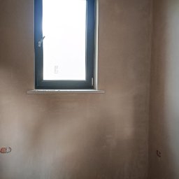 AMBITION Jarosław Rozwadowski - Pokój po nałożeniu tynku, widoczne ślady po instalacjach elektrycznych, okno z czarną ramą i drewniane belki stropowe. Ściany w kolorze beżowym.
