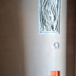 AMBITION Jarosław Rozwadowski - Ściana po tynkowaniu z zamontowaną skrzynką elektryczną i puszkami pod gniazdka. Widoczne przewody elektryczne. Prace wykończeniowe w trakcie realizacji.