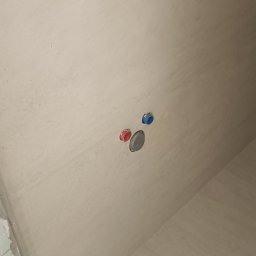 AMBITION Jarosław Rozwadowski - Ściana po nałożeniu gładzi, widoczne wyprowadzenia instalacji wodnej z zaślepkami w kolorze niebieskim i czerwonym oraz otwór kanalizacyjny.