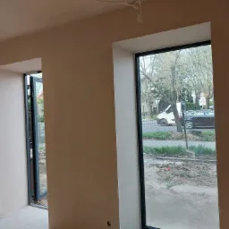 Świeżo otynkowane wnętrze z widocznymi oknami i drzwiami balkonowymi, drabina malarska oparta o ścianę w lewym dolnym rogu, surowa żarówka zwisająca z sufitu.