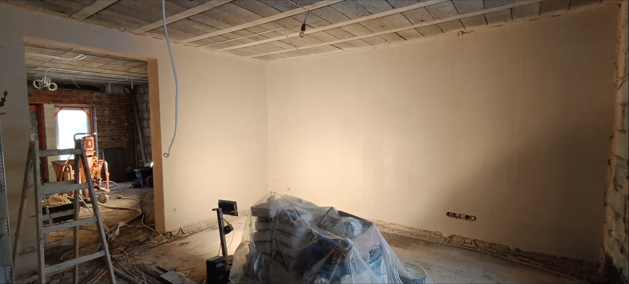 Świeżo otynkowane ściany w pomieszczeniu w trakcie remontu, widoczne worki z zaprawą pod folią, drabina i agregat tynkarski w tle.