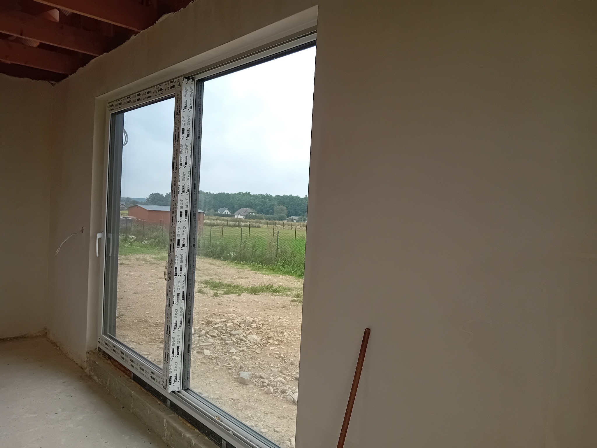 Świeżo otynkowane wnętrze z zamontowanym oknem przesuwnym, widok na zewnątrz na wiejski krajobraz, surowy stan wykończenia podłogi i sufitu.
