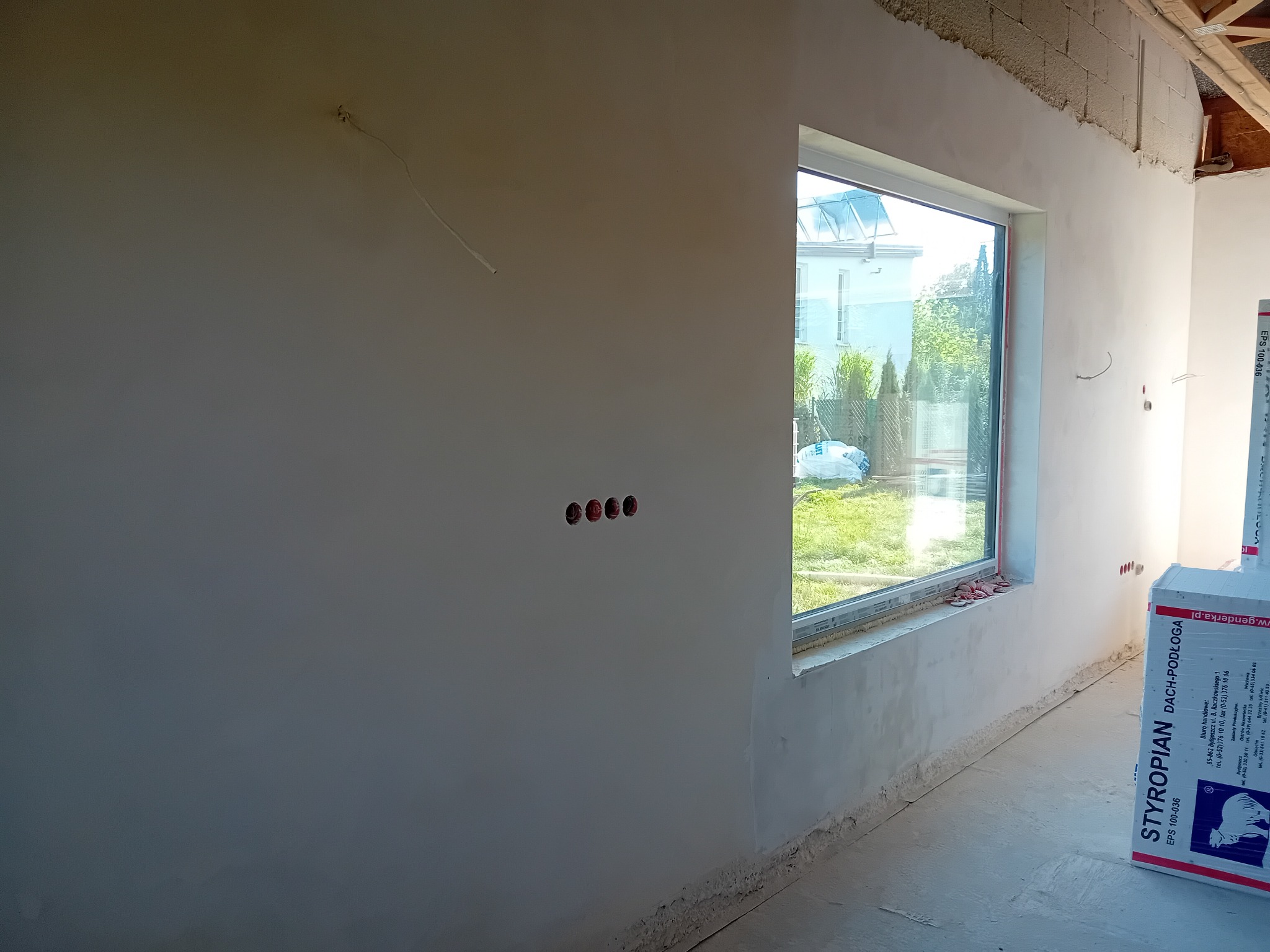 Świeżo otynkowana ściana wewnętrzna z widocznymi puszkami elektrycznymi i oknem, częściowo zakryta paczką styropianu podłogowego.