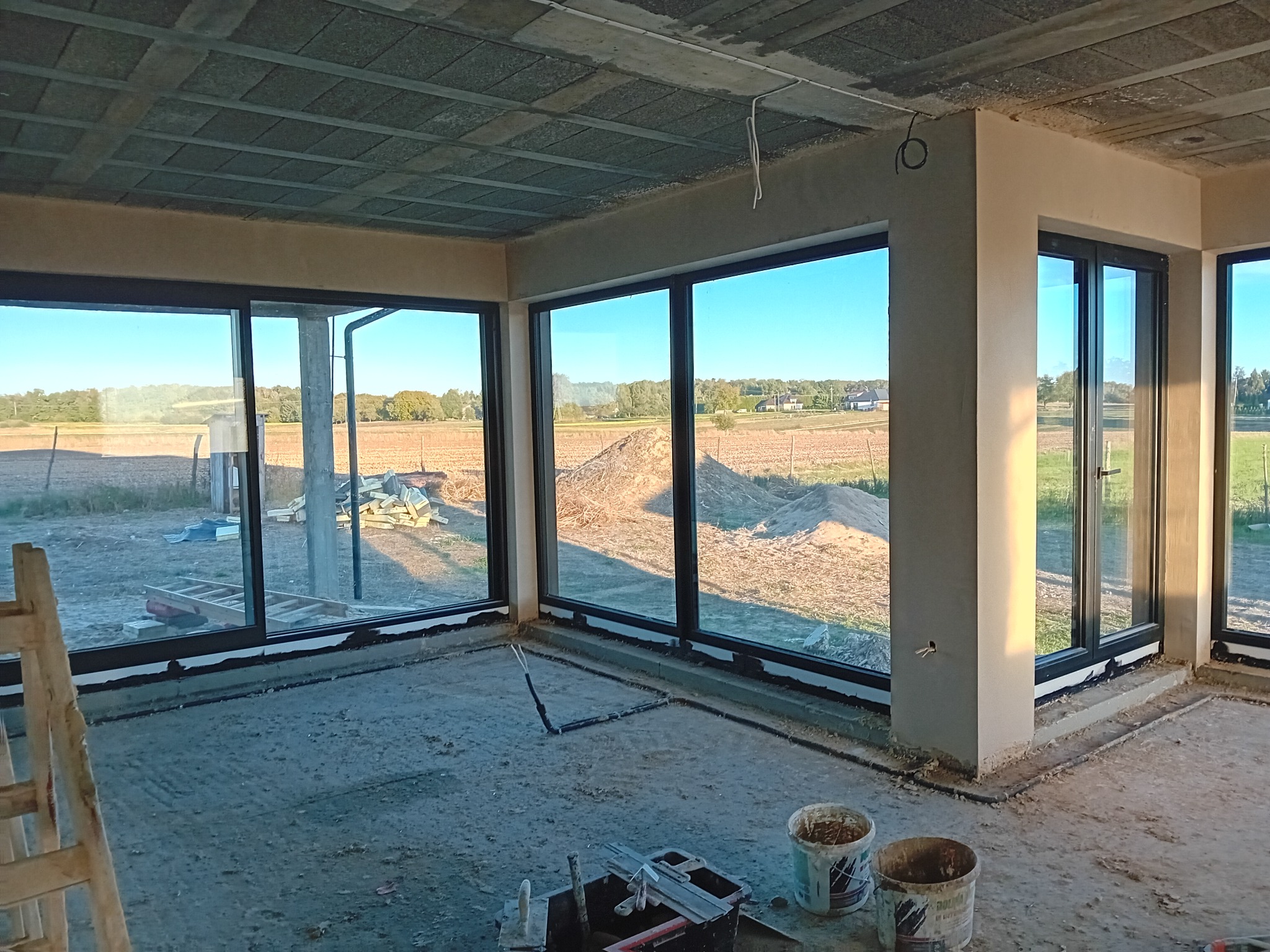 Wnętrze budynku w stanie surowym zamkniętym po tynkowaniu ścian, z dużymi oknami z widokiem na pole, widoczne narzędzia i wiaderka po farbie.