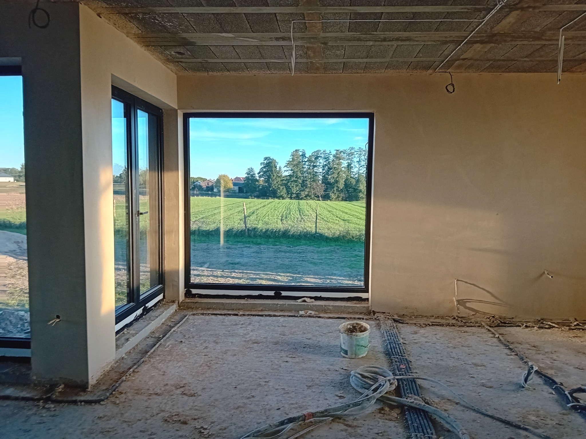 Wnętrze surowego domu po nałożeniu tynku na ściany, z widokiem na zielone pole przez duże okno. Widoczne kable elektryczne na podłodze i wiadro po zaprawie.