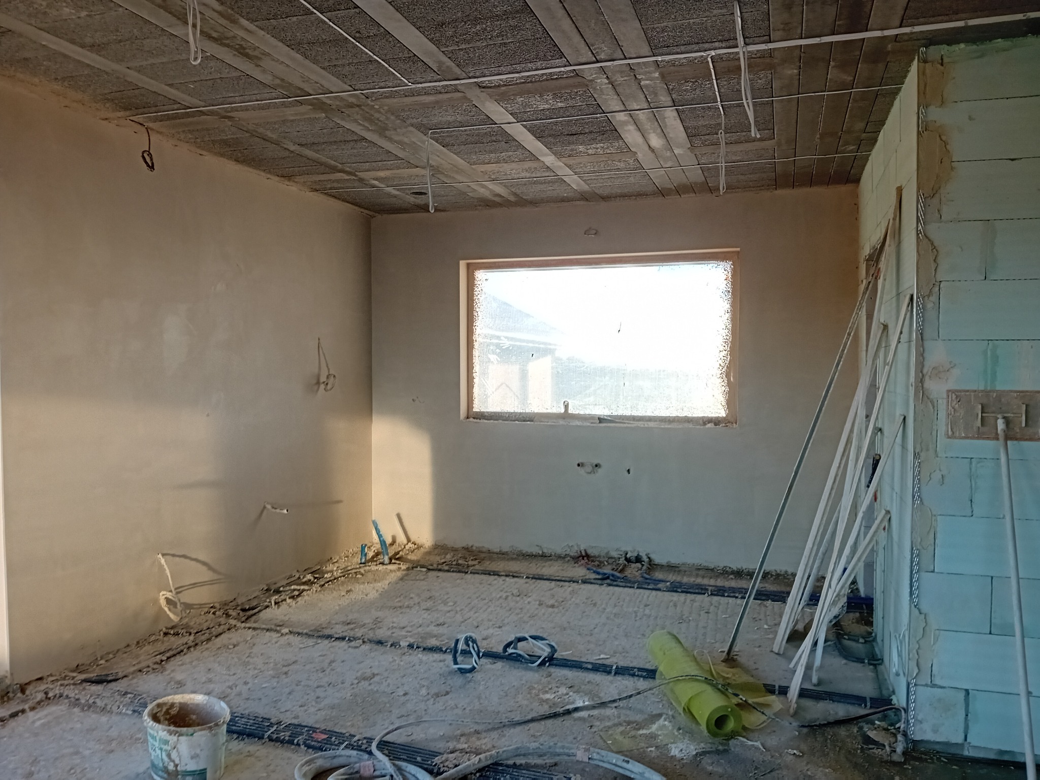 Wnętrze w trakcie prac wykończeniowych: ściany po nałożeniu pierwszej warstwy tynku, widoczne instalacje podłogowe, profile do tynkowania przy ścianie, sufit z betonowych płyt.
