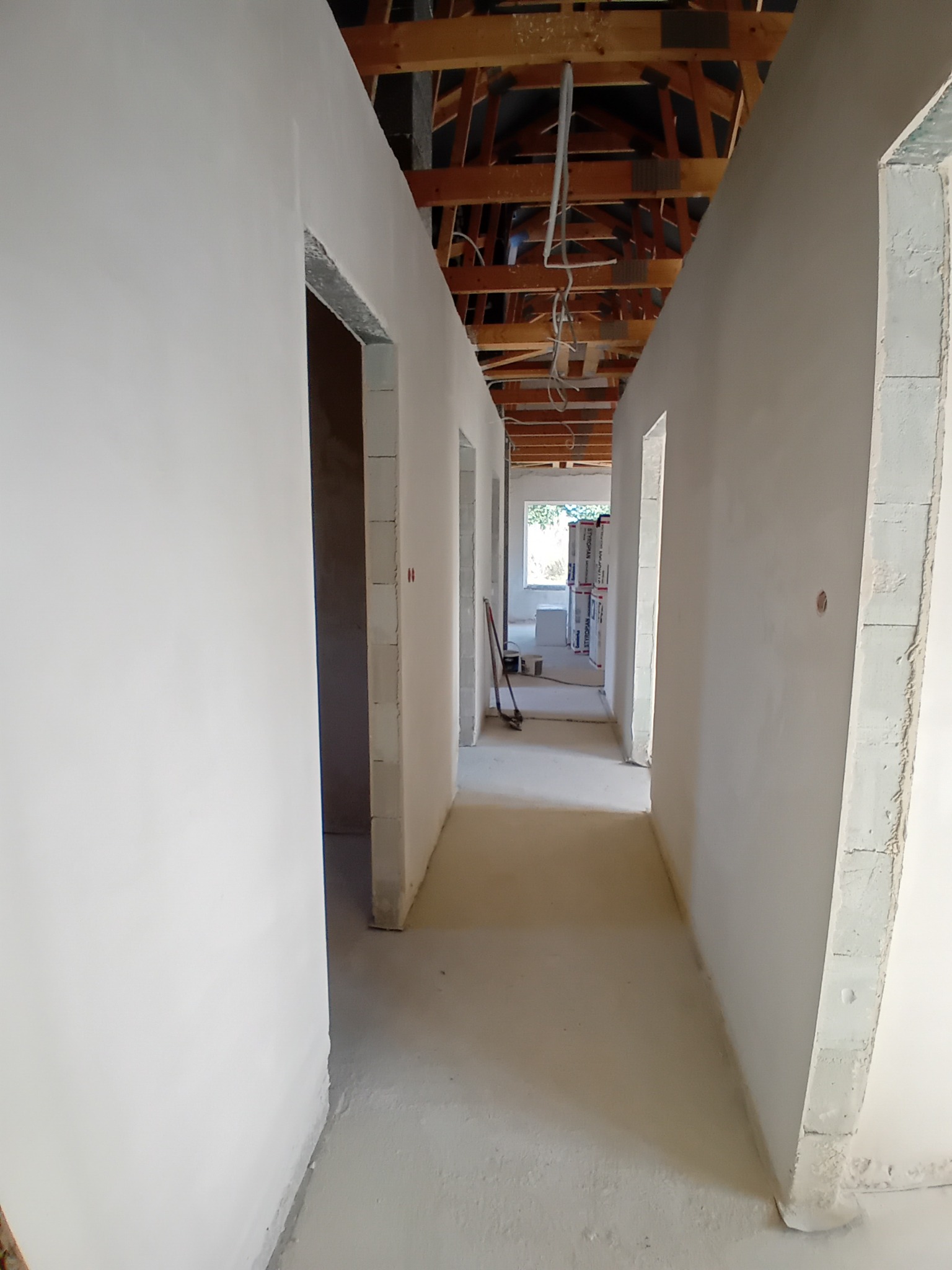Wnętrze w trakcie prac wykończeniowych: długi korytarz ze świeżo otynkowanymi ścianami, widoczne drewniane elementy konstrukcji dachu, w tle materiały budowlane.