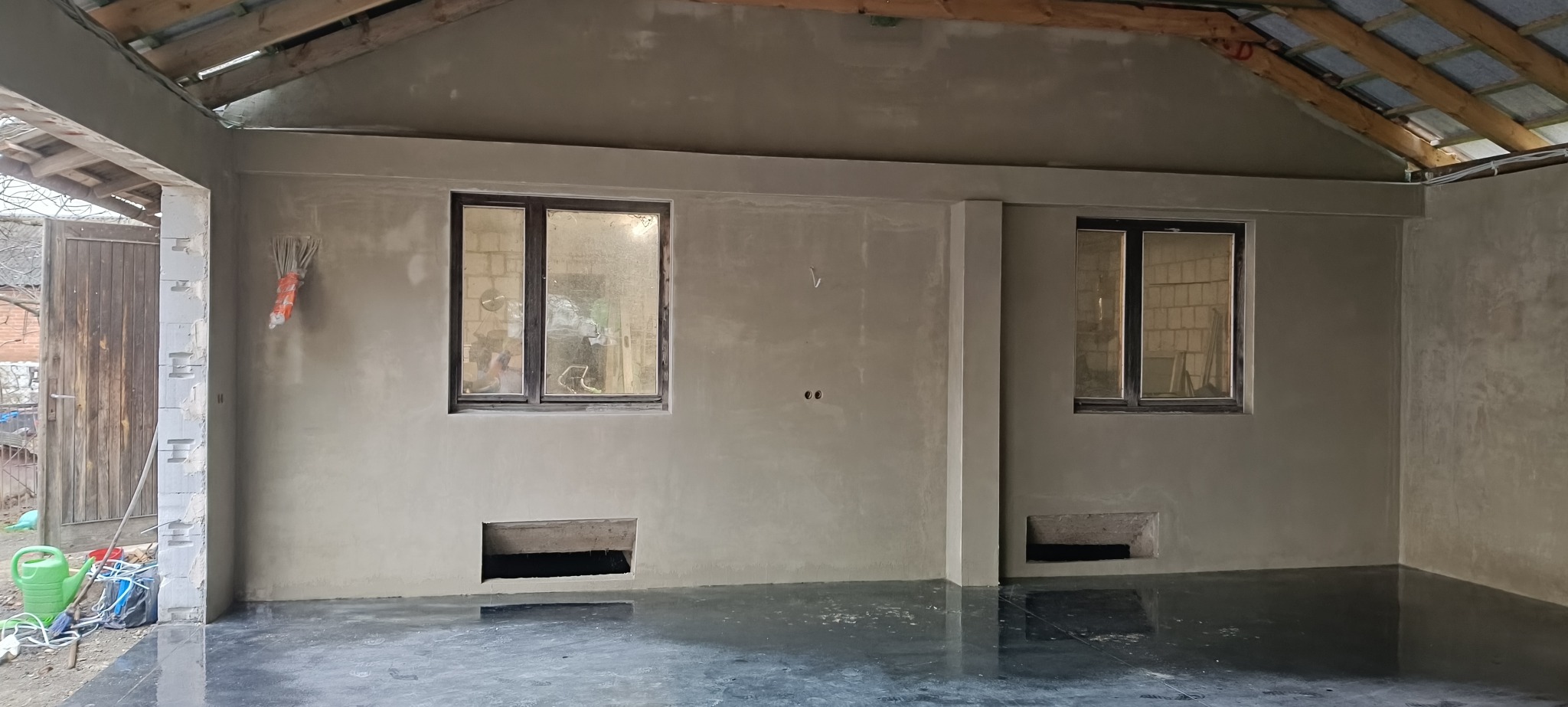 Wnętrze po tynkowaniu: ściany pokryte świeżym tynkiem, widoczne okna i otwory w ścianach. Podłoga mokra, odbijająca światło. Surowy stan wykończenia.