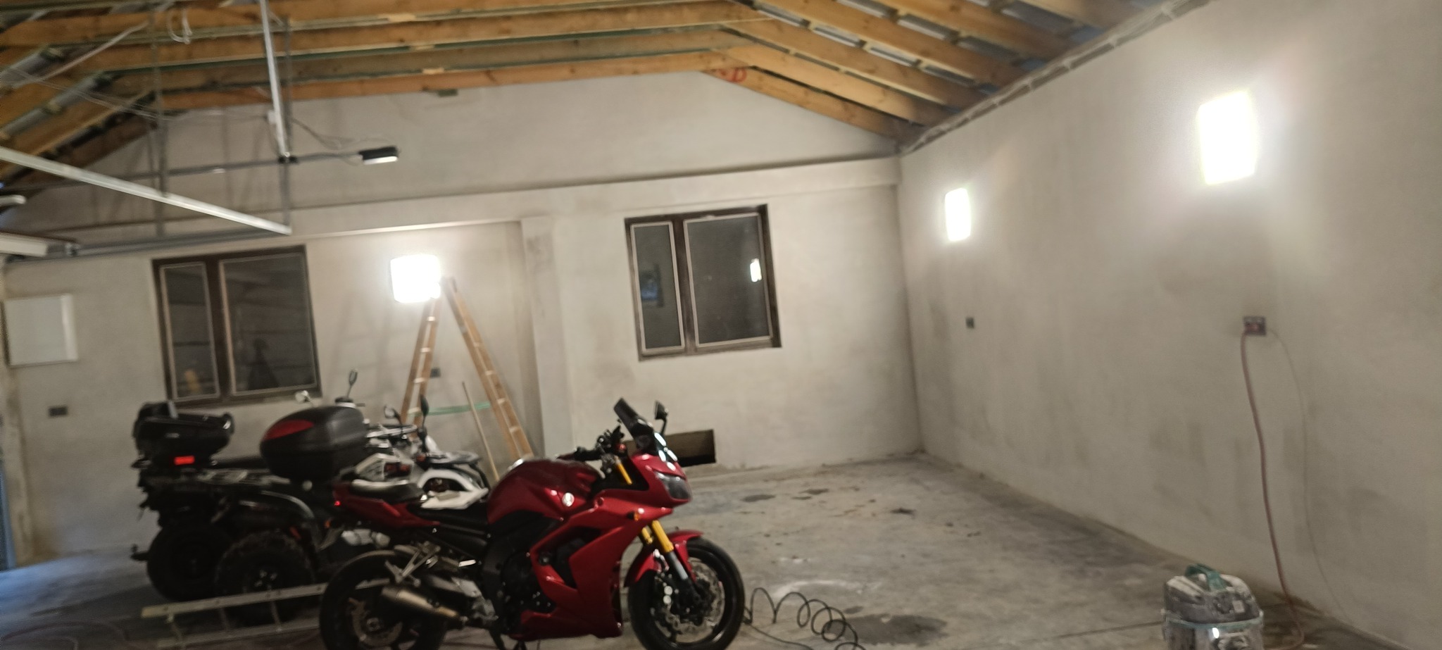 Wnętrze garażu po szpachlowaniu ścian, z widocznymi oknami, drewnianą konstrukcją dachu i motocyklami. Ściany przygotowane do dalszych prac wykończeniowych.