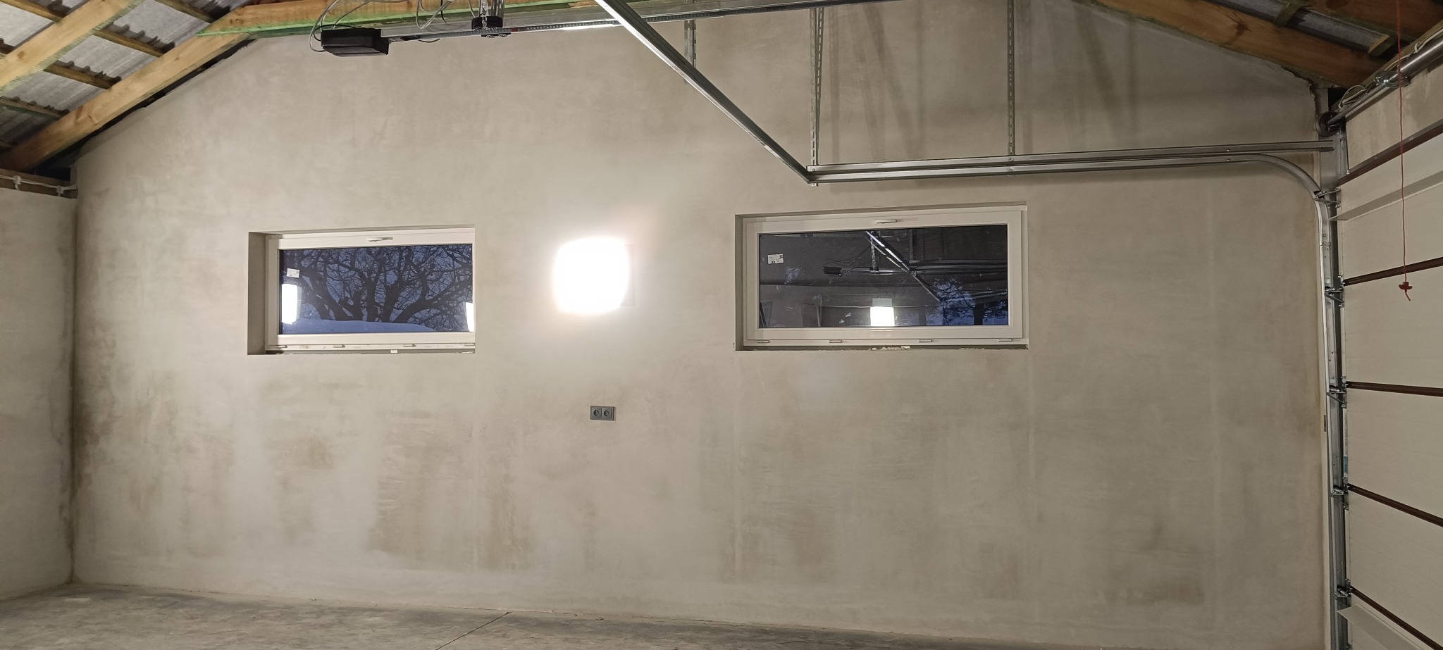 Ściana garażu po nałożeniu tynku, widoczne dwa okna z odbiciem drzew, surowy beton na podłodze, szyny do bramy garażowej.