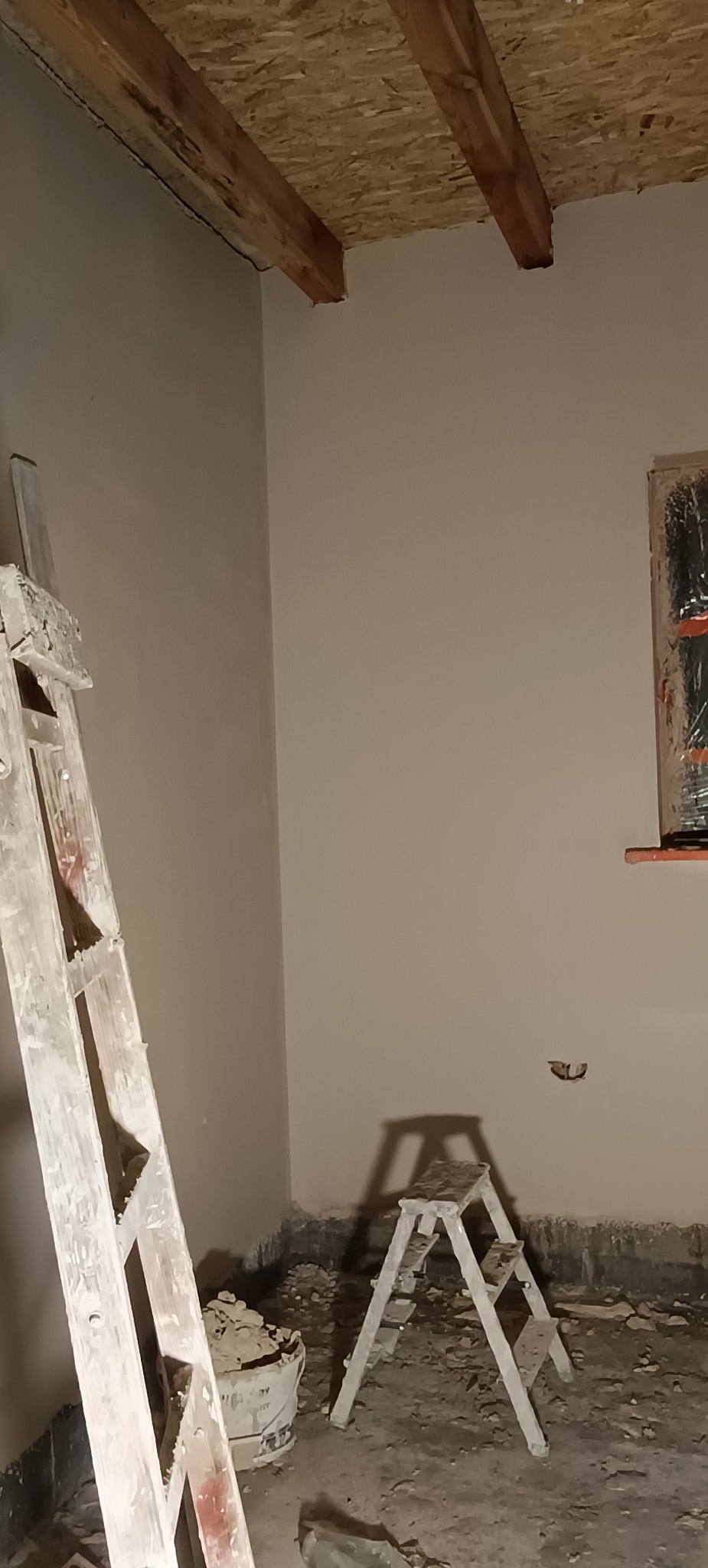 Świeżo pomalowane ściany w pomieszczeniu z drewnianym stropem, drabina malarska i schodki, widoczne ślady prac wykończeniowych na podłodze.