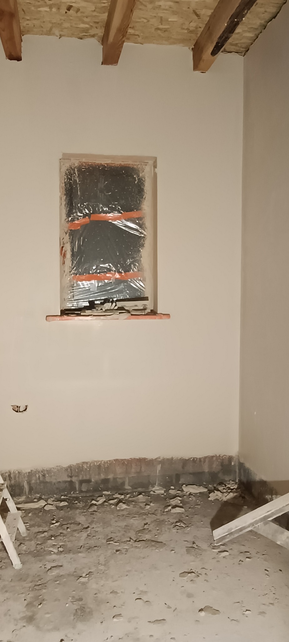 Przygotowane okno do prac wykończeniowych, zabezpieczone folią. Ściany po szpachlowaniu, surowy strop z drewnianych belek, podłoga w trakcie prac remontowych.