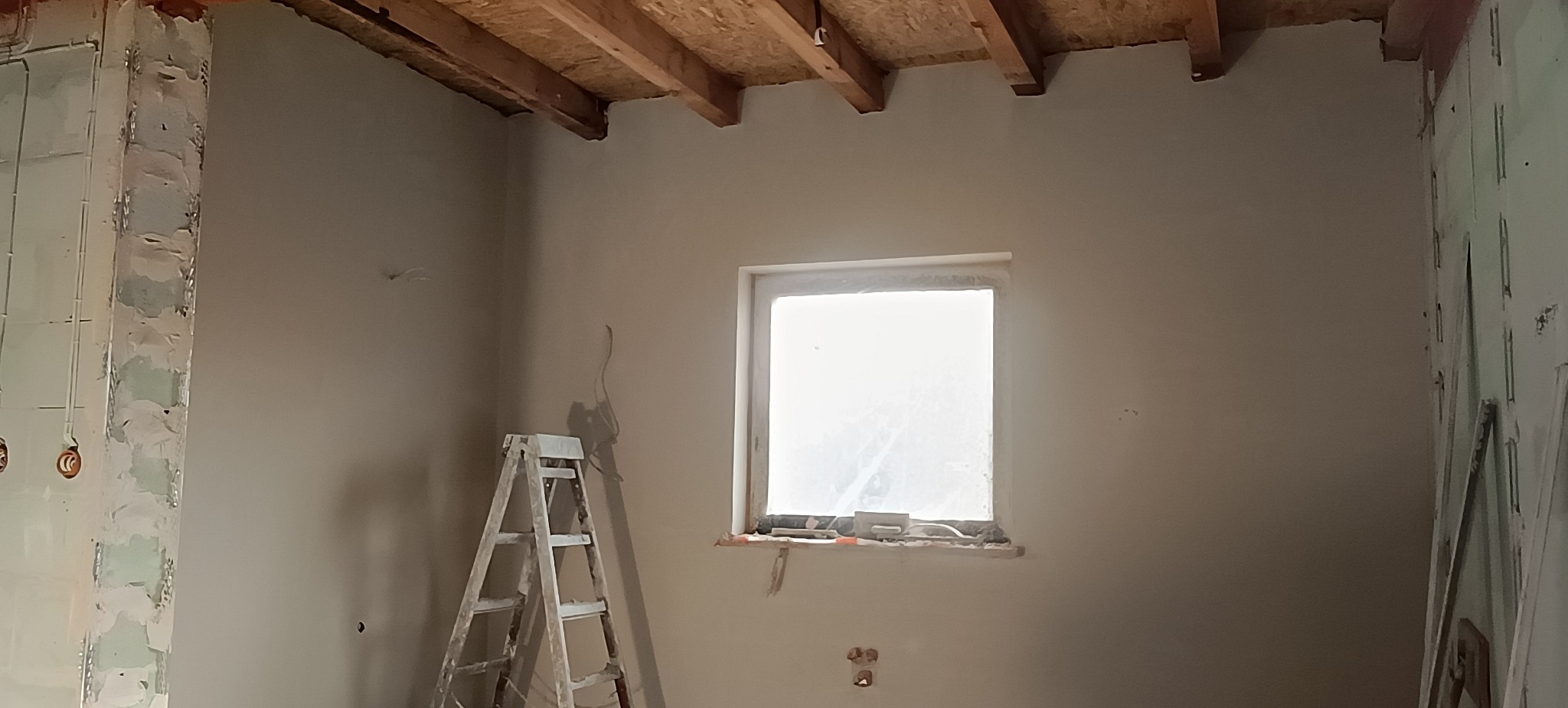 Wnętrze po pracach przygotowawczych w Garbowie: ściany po nałożeniu gładzi, widoczna drabina malarska i surowy strop. Okno zabezpieczone folią.
